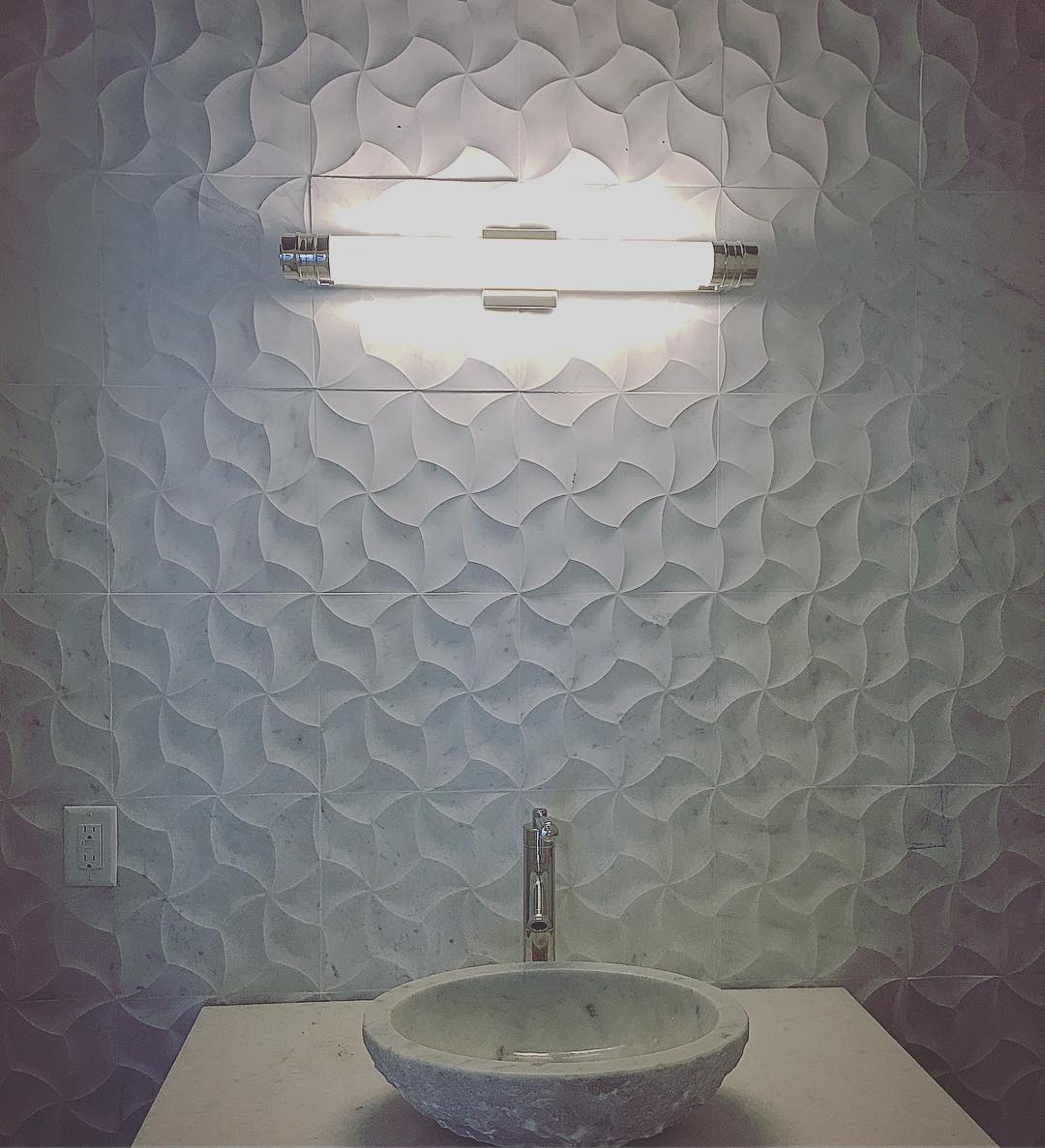 Bathroom vanity with wavy textured white marble tile backsplash, stone vessel sink, and illuminated modern light bar.